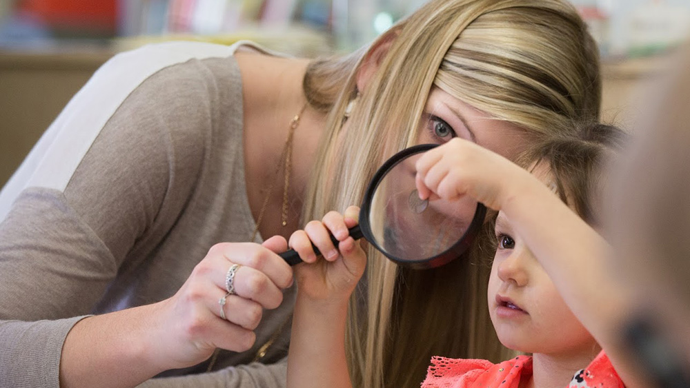 An instructor helps a young child hold a magnifying glass.