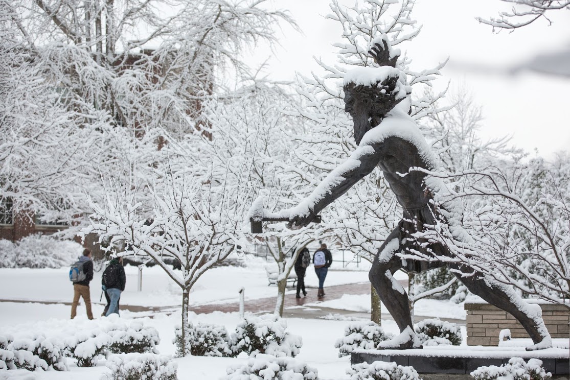 A statue of PennWest California's mascot, the Vulcan, stands snow-covered as students walk past to get to class.