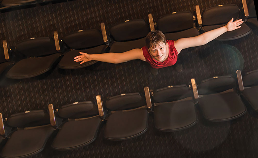 Alyssa Freeman looks upward with her arms stretched outwards in the aisle of theatre seating.